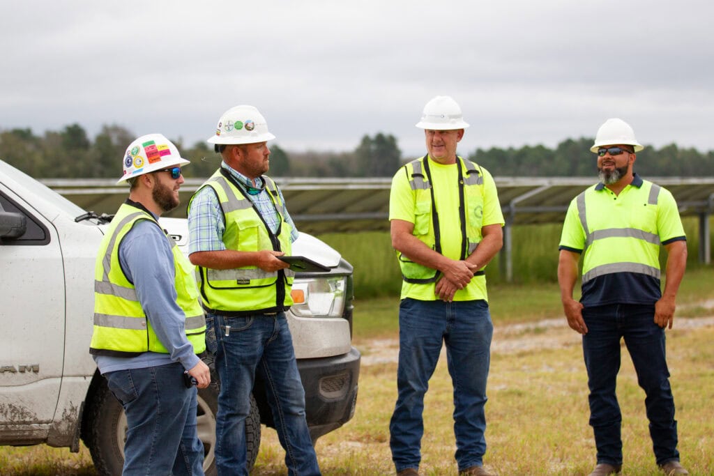 A group of men in safety vests and hard hats stand near a truck talking