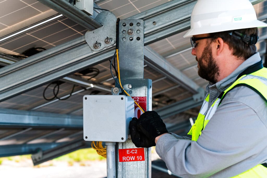 A man in a hard hat looks at wires under a solar panel