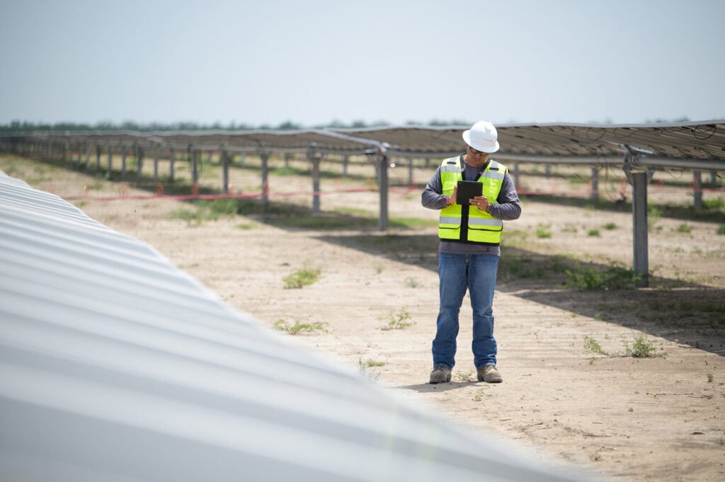 A man in safety gear writes something on a tablet between two rows of solar panels